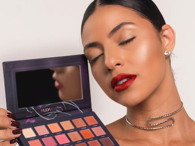Beautiful brunette woman with bold lipstick holding an eyeshadow palette against a white studio background.