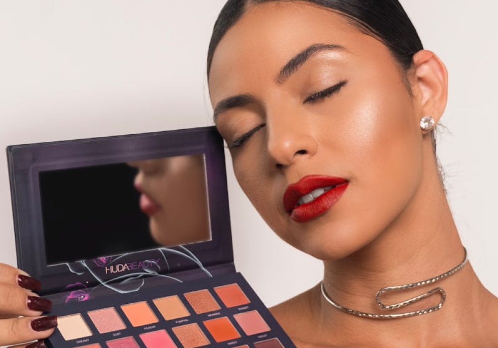 Beautiful brunette woman with bold lipstick holding an eyeshadow palette against a white studio background.