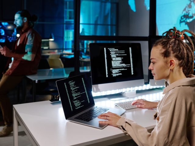 Serious female programmer with dreadlocks decoding data on laptop screen while sitting by desk with computer monitor in dark office