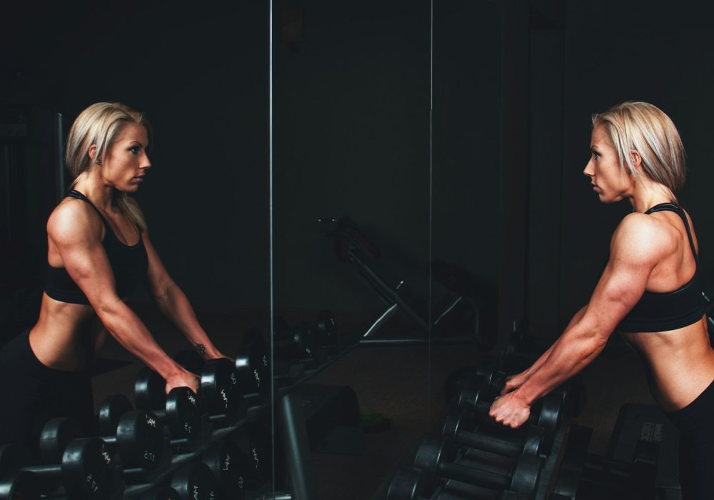 woman wearing black top top holding black dumbbells standing in front of mirror