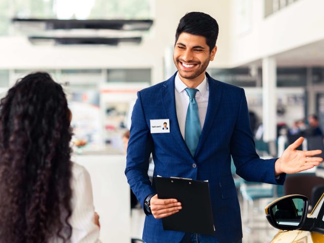 Wealthy middle-eastern young woman choosing new car, having conversation with friendly sales manager at brand new vip showroom, dealer pointing at yellow comfy auto and smiling, side view